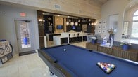 This single-floor space is designed for upscale entertaining, combining a games area, bar, and lounge into one cohesive, open-plan layout. At the forefront sits a full-size pool table with a striking blue felt surface, positioned on light travertine-style tile flooring that extends throughout the space.

The built-in bar area serves as the architectural centerpiece, featuring dark cabinetry with open display shelving, a large white waterfall-edge counter, and tall bar seating. A series of globe pendant lights hang above the bar, casting warm ambient light and adding a contemporary design element. Wall sconces throughout contribute to the layered lighting scheme, enhancing the overall atmosphere.

Wood-paneled ceilings in a light natural finish run across the space, adding warmth and texture while complementing the contrast between the dark cabinetry and light surfaces. A built-in media unit with a mounted television is integrated into the bar area. Adjacent to the games zone, a comfortable lounge seating area provides a relaxed retreat, while framed wall art adds a curated finishing touch to this well-appointed entertainment space.