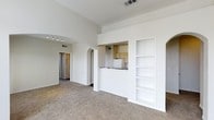 This single-floor space showcases an open-concept layout that promotes a sense of spaciousness throughout. The living area is finished with wall-to-wall carpet and benefits from notably tall, vaulted ceilings that amplify the feeling of openness and airiness. Crisp white walls and trim create a clean, neutral backdrop throughout the space.

A built-in shelving unit is recessed into one wall of the living area, offering practical display and storage options. An arched passageway leads from the living space into a hallway, where track lighting is mounted overhead, adding a functional decorative element. The hallway connects to additional rooms, suggesting a well-considered floor plan.

The kitchen is positioned adjacent to the living area and is accessible via a pass-through counter with a dark countertop, allowing visual connection between the two spaces. White cabinetry and white appliances are visible within the kitchen, maintaining the consistent light and neutral aesthetic found throughout the unit. A ceiling vent indicates a central HVAC system. Overall, the space presents a straightforward, functional design with architectural details that add character.