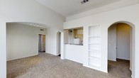 This single-floor space showcases an open-concept layout that promotes a sense of spaciousness throughout. The living area is finished with wall-to-wall carpet and benefits from notably tall, vaulted ceilings that amplify the feeling of openness and airiness. Crisp white walls and trim create a clean, neutral backdrop throughout the space.

A built-in shelving unit is recessed into one wall of the living area, offering practical display and storage options. An arched passageway leads from the living space into a hallway, where track lighting is mounted overhead, adding a functional decorative element. The hallway connects to additional rooms, suggesting a well-considered floor plan.

The kitchen is positioned adjacent to the living area and is accessible via a pass-through counter with a dark countertop, allowing visual connection between the two spaces. White cabinetry and white appliances are visible within the kitchen, maintaining the consistent light and neutral aesthetic found throughout the unit. A ceiling vent indicates a central HVAC system. Overall, the space presents a straightforward, functional design with architectural details that add character.