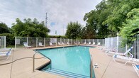 This outdoor pool area offers a well-maintained and inviting aquatic retreat. The generously sized rectangular swimming pool features clear, light-blue water with clearly marked depth indicators along its edges, ensuring safety for all users. The pool is finished with classic tile detailing and provides easy water access via stainless steel entry rails.

Surrounding the pool is a broad concrete deck that offers ample space for relaxation and movement. Numerous lounge chairs and seating options are arranged along the poolside, providing plenty of room for guests to unwind. A white metal fence encloses the entire perimeter of the pool area, offering both security and a clean, classic aesthetic.

The backdrop of the space is framed by mature, leafy trees that provide a natural privacy screen and a touch of greenery, enhancing the outdoor ambiance. The open-sky setting allows for abundant natural light throughout the day. Informational signage is posted within the enclosure, consistent with a well-organized and safety-conscious facility. Overall, the pool area presents a clean, functional, and pleasant environment well-suited for leisure and recreational use.