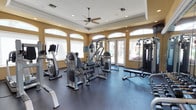 This fitness center offers a thoughtfully designed workout environment that balances functionality with an inviting aesthetic. The space features a tray ceiling with crown molding and a central ceiling fan, contributing to both the refined architectural character and comfortable air circulation during workouts. Warm golden-yellow walls complement the dark rubber flooring, which provides durability and traction underfoot.

Natural light floods the room through a series of arched windows along the walls, as well as French doors that open to an outdoor area with landscaped greenery and flowering plants visible beyond. Recessed lighting supplements the natural light, ensuring the space remains bright at all hours.

The fitness center is outfitted with a comprehensive selection of equipment, including stationary bicycles, elliptical trainers, multi-station cable and weight machines, a weight bench, and additional strength training apparatus. The layout is open and spacious, allowing for comfortable movement between stations. The combination of cardio and resistance training equipment makes this a versatile facility suited to a wide range of fitness routines. Overall, the space strikes an effective balance between a polished, welcoming design and a fully functional workout environment.