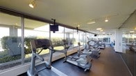 This fitness center offers a clean, functional workout environment designed to support a comprehensive exercise routine. The space is lined with floor-to-ceiling windows along one wall, flooding the interior with abundant natural light and providing open views of the surrounding landscaped greenery — creating an inviting atmosphere for exercise.

The cardio area is well-appointed with a row of modern Life Fitness machines, including treadmills, ellipticals, and stationary bikes, all positioned to take full advantage of the garden views. Overhead, ceiling-mounted flat-screen televisions provide entertainment options for users during their workouts.

Track lighting and recessed ceiling fixtures provide ample illumination throughout the space, complementing the natural light from the expansive windows. The flooring features a durable, dark rubberized surface well-suited for high-traffic fitness use. The ceiling has a textured finish and maintains a comfortable height, contributing to the open, airy feel of the room.

Overall, the fitness center strikes a balance between practicality and comfort, offering a bright, organized, and fully equipped environment ideal for daily cardio training. The seamless connection to the outdoors through the large windows enhances the overall ambiance, making workouts a more pleasant experience.