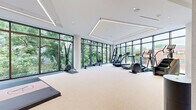 Located at 4909 Auburn Ave in Bethesda, MD, this professionally designed fitness center offers a comprehensive and upscale wellness environment across a generously sized single-floor layout. The main exercise room is a standout feature, characterized by floor-to-ceiling windows along two walls that flood the space with natural light and frame lush, verdant outdoor views. Linear LED ceiling fixtures complement the natural light, lending the space a clean, modern aesthetic. The room accommodates a full array of cardio equipment — including treadmills, ellipticals, rowing machines, and a stair climber — alongside dedicated free weight and strength training zones featuring Eleiko and Rogue equipment, kettlebells, dumbbells, and resistance accessories. A mirrored wall-mounted display and framed artwork add visual interest. A separate glass-enclosed studio, clearly marked and accessible via sleek black-framed doors with full mirror paneling, provides space for classes or individual training. The hallways connecting the facility are bright and refined, with light tile flooring, warm wood ceiling accents, and living plant installations that bring a natural, welcoming touch. A stylish stairwell area features rich wood detailing and glass railings, reinforcing the facility's high-end character throughout.