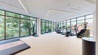 Located at 4909 Auburn Ave in Bethesda, MD, this professionally designed fitness center offers a comprehensive and upscale wellness environment across a generously sized single-floor layout. The main exercise room is a standout feature, characterized by floor-to-ceiling windows along two walls that flood the space with natural light and frame lush, verdant outdoor views. Linear LED ceiling fixtures complement the natural light, lending the space a clean, modern aesthetic. The room accommodates a full array of cardio equipment — including treadmills, ellipticals, rowing machines, and a stair climber — alongside dedicated free weight and strength training zones featuring Eleiko and Rogue equipment, kettlebells, dumbbells, and resistance accessories. A mirrored wall-mounted display and framed artwork add visual interest. A separate glass-enclosed studio, clearly marked and accessible via sleek black-framed doors with full mirror paneling, provides space for classes or individual training. The hallways connecting the facility are bright and refined, with light tile flooring, warm wood ceiling accents, and living plant installations that bring a natural, welcoming touch. A stylish stairwell area features rich wood detailing and glass railings, reinforcing the facility's high-end character throughout.
