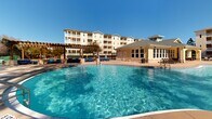 Located at 7381 Navarre Pkwy in Navarre, Florida, this outdoor amenity space presents a well-appointed resort-style pool area designed for relaxation and recreation. The centerpiece of the space is a freeform, curved swimming pool with crystal-clear blue water and gentle fountain features, complemented by a second, more rectangular pool nearby — both offering ample room for leisure. The pool deck is finished with attractive brick pavers and is generously lined with lounge chairs and seating areas, providing plenty of space to unwind under the Florida sun. A charming pergola structure adds architectural interest and a shaded gathering spot alongside the pools. Adjacent to the pool area stands a single-story clubhouse building with large windows, a covered porch, and warm yellow exterior tones that complement the surrounding residential buildings. The multi-story residential structures visible in the background feature balconies and neutral coastal-inspired facades. The entire pool area is enclosed by a wrought iron fence and is beautifully framed by tall, mature pine trees that lend a natural, serene ambiance. The combination of thoughtful landscaping, quality amenities, and abundant natural light creates an inviting outdoor retreat ideal for community living in the Navarre, FL area.