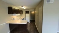 This single-story space centers around a well-organized kitchen that combines practicality with a clean, modern aesthetic. The kitchen is outfitted with dark espresso-toned cabinetry that extends to the upper walls, providing ample storage. White appliances, including a freestanding electric range, dishwasher, and refrigerator, create a crisp contrast against the darker cabinetry. The countertops are finished in a light, neutral tone, adding brightness to the space. An under-cabinet lighting system illuminates the work surfaces effectively. A peninsula-style counter extension provides additional prep space and defines the kitchen area. The flooring throughout is a rich, dark hardwood-style material that runs continuously from the kitchen into the adjacent hallway, contributing to a cohesive feel. A ceiling-mounted fluorescent light fixture provides overhead illumination. A central HVAC vent is visible in the ceiling, indicating a climate control system. The hallway beyond the kitchen leads to additional areas of the home, suggesting a practical, linear layout. Neutral-toned walls throughout keep the space feeling clean and versatile.