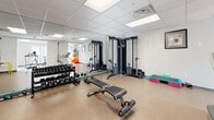 Located at 700 King St W, this fitness facility offers a thoughtfully designed workout space spread across a single open floor. The room features a clean, modern aesthetic with white walls, a drop tile ceiling with integrated panel lighting, and light hardwood-style flooring that lends a bright and welcoming atmosphere throughout.

The space is divided into distinct functional zones. The strength training area is well-appointed with a full dumbbell rack, a multi-station cable machine, an adjustable weight bench, and a selection of medicine balls and step platforms. Full-length mirrors line portions of the walls, enhancing the sense of openness and allowing users to monitor their form during exercise.

The cardio zone is equipped with a treadmill, elliptical trainer, and stationary bike, positioned near a windowed area that allows natural light to filter in, creating an energizing environment. Suspension training straps (TRX-style) and vibrant yellow parallel bars provide additional options for functional and bodyweight training.

Open floor space throughout the facility supports stretching, floor exercises, and group activity. The layout is efficient and uncluttered, maximizing usability. A waste station and wall-mounted informational panels contribute to the facility's clean and organized presentation. Access is provided via a secure door entry.
