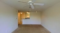 This single-floor space showcases an open-concept layout that seamlessly connects the living area to the kitchen. The living room is finished with wall-to-wall carpeting in a warm neutral tone, creating a comfortable and cohesive feel underfoot. A white ceiling fan with a pull-chain light fixture is centrally mounted, providing both air circulation and ambient lighting to the space.

The kitchen is positioned at the rear of the main living area and features a raised pass-through counter that serves as a natural divider between the two spaces while maintaining an open, connected feel. The kitchen is equipped with overhead cabinetry in a light finish and includes standard appliances, illuminated by dedicated overhead lighting. The pass-through design allows for easy interaction between the kitchen and living areas.

The entryway is visible to the left, framed by a standard doorway with warm lighting, offering a welcoming transition into the unit. The walls throughout are painted in a clean, neutral off-white, contributing to a bright and versatile atmosphere. The overall design prioritizes functionality and simplicity, making the space highly adaptable to a variety of furnishing styles.