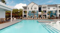 Located at 347 Arboretum Dr in Wilmington, North Carolina, this outdoor amenity space offers a resort-inspired poolside experience designed for relaxation and socializing. The centerpiece is a generously sized swimming pool with crystal-clear blue water, complemented by a spacious concrete sundeck lined with lounge chairs and umbrellas for sun-soaking comfort. Rows of chaise lounges border the pool, providing ample space for residents to unwind under the coastal Carolina sky. Vibrant teal accent chairs and small bistro tables are thoughtfully arranged along the pool's edge, adding a pop of color against the neutral hardscape. A gated entry with stone tile flooring provides a seamless transition between the covered patio and the pool deck. The covered outdoor patio area features elegant grey stone tile flooring, wicker bar seating, and large glass windows that connect the interior clubhouse to the outdoors. A warm wooden pergola and a dedicated pool house structure offer shaded retreats and additional gathering space. Tropical palm trees punctuate the landscape, reinforcing the coastal aesthetic, while the multi-story residential buildings clad in bold blue and white board-and-batten siding frame the backdrop. The overall ambiance is vibrant, polished, and inviting, with a strong sense of community and coastal charm.