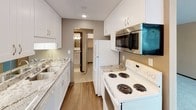 This single-floor unit offers a functional and comfortable layout with a neutral color palette of warm beige tones throughout. The carpeted living and bedroom areas are bright and open, each featuring windows with horizontal blinds that allow natural light to filter in and provide views of surrounding greenery. Baseboard heating units run along the walls, and a wall-mounted air conditioning unit ensures year-round climate comfort. The living area is generously sized, offering ample room for furnishings, and includes a closet with classic bifold doors as well as a solid wood entry door. The kitchen has been thoughtfully updated with crisp white cabinetry, granite-look countertops, and a stainless steel over-the-range microwave, complemented by a white electric range and refrigerator. Wood-look flooring adds warmth to the kitchen, which connects directly to the bathroom and additional spaces beyond. The bathroom is clean and well-lit, featuring a tiled tub-and-shower combination, white fixtures, a granite-topped vanity with an undermount sink, and a large mirror with a multi-bulb light bar. Textured ceilings are present throughout the unit, adding a classic residential character to the overall space.
