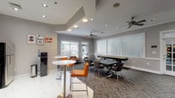 This single-floor community amenity space is designed to serve as a versatile social and recreational hub. The main gathering area is finished with textured herringbone-patterned carpet and neutral gray walls accented by white wainscoting, creating a clean and modern aesthetic throughout. Ceiling fans are positioned throughout the room to maintain airflow and comfort.

The space is furnished with a large black dining-style table surrounded by contemporary chairs, ideal for group gatherings or casual meetings. High-top bistro tables with vibrant orange bar stools provide an additional seating option, while leather lounge chairs and side tables offer a relaxed retreat. A full-length shuffleboard table runs along one wall, serving as a central recreational feature. Wall-mounted flat-screen TVs are placed at multiple points around the room for entertainment. Large windows and glass-paned doors allow natural light to filter in and offer views of the surrounding outdoors.

A dedicated kitchenette area includes gray cabinetry with dark granite countertops, a single basin sink, a commercial-style coffee machine, and a compact refrigerator — all positioned beneath a wall-mounted TV. Frosted glass panel dividers provide a subtle visual boundary within the space without compromising its open, communal feel. Recessed lighting complements the natural light to keep the entire space bright and inviting.