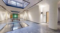 This single-floor gallery space offers an open, adaptable layout designed to accommodate a wide range of exhibitions and events. The architectural design draws immediate attention with its large overhead skylights that flood the interior with abundant natural light, creating a bright and airy atmosphere throughout. Exposed structural columns with rounded bases punctuate the floor plan, adding character while defining distinct zones within the open layout. Polished concrete floors extend across the entire space, lending an industrial yet refined aesthetic. Overhead, adjustable track lighting runs along black rails mounted to the ceiling, providing flexible, directed illumination ideal for showcasing artwork or displays. A notable feature of the floor is a recessed glass panel inset, which allows light to pass through and adds a distinctive architectural detail. The walls are finished in crisp white, offering expansive, uninterrupted display surfaces, while a bold green accent wall anchors one end of the space, providing visual contrast. Exposed piping and raw ceiling finishes reinforce the industrial character of the interior. A wooden bench with a glass top serves as a central furnishing element. Additional utility doors are accessible at the perimeter, and wall-mounted sconce lighting supplements the track system. Overall, the space balances raw industrial elements with clean, gallery-quality finishes.