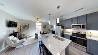 This 1-bedroom unit showcases a thoughtfully designed open-concept layout that integrates the kitchen, dining, and living areas into one cohesive, light-filled space. The kitchen is a standout feature, anchored by a large granite-topped island with a double undermount sink, bar seating, and pendant lighting overhead. Dark navy cabinetry contrasts elegantly with the light stone countertops, while stainless steel appliances — including a refrigerator, electric range, and built-in microwave — add a polished, modern touch. A subway tile backsplash completes the kitchen's clean aesthetic. The living area extends naturally from the kitchen, featuring warm wood-look flooring, a ceiling fan, and a bright window with access to what appears to be an outdoor balcony, flooding the space with natural light. The open dining area connects both zones seamlessly. Throughout the unit, the neutral wall tones, contemporary light fixtures, and cohesive design elements create a modern and inviting atmosphere well-suited for comfortable everyday living.