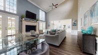 This well-appointed single-floor space combines elegance and functionality across several distinct areas. The centerpiece is a grand lounge featuring soaring vaulted ceilings with crown molding, rich hardwood-style flooring, and a striking fireplace with an ornate cream-colored surround topped by a wall-mounted television. Tall multi-pane windows flood the room with natural light, while a ceiling fan adds to the airy atmosphere. Seating is arranged thoughtfully throughout, including a large sectional sofa in warm grey accented with teal throw pillows, a glass-top coffee table, and patterned accent chairs. Wainscoting along the lower walls adds a refined architectural detail throughout the space. An adjacent sitting area features bold teal wingback chairs, a console table, and vibrant contemporary artwork, creating an inviting transitional zone. The kitchenette is equipped with white raised-panel cabinetry, granite-look countertops, a textured stone tile backsplash, and stainless steel appliances including a side-by-side refrigerator and dishwasher. A dedicated workstation room offers a long floating desk with multiple computer terminals, complemented by a slate-blue accent wall and modern bar-height seating. The cohesive teal, grey, and neutral color palette throughout gives the entire space a polished, contemporary feel.