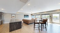 Located at 240 Thompson Ave E in West St. Paul, MN, this contemporary amenity space offers a well-appointed, single-floor layout designed for both social gatherings and relaxed lounging. The interior lounge area features light wood-look flooring, tray ceilings with recessed lighting, and large floor-to-ceiling windows and sliding glass doors that flood the space with natural light while providing direct access to the outdoor terrace. Thoughtfully curated seating arrangements include plush sofas, leather accent chairs, and high-top bar tables, complemented by a striking black marble-topped bar counter and a bold geometric pendant light fixture. A wall-mounted flat-screen TV anchors the living area, adding a functional entertainment element. The kitchenette is outfitted with dark wood cabinetry, quartz countertops featuring marble-like veining, stainless steel appliances, and a decorative hexagonal tile backsplash. The expansive outdoor rooftop terrace is a standout feature, clad in large-format pavers and enclosed by vibrant teal panel walls. It offers abundant seating with sectional sofas, dining tables, and lounge chairs, along with a built-in grill area and overhead string lights that create an inviting atmosphere day or night.