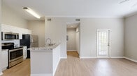 Located at 5010 Central Sarasota Pkwy in Sarasota, FL, this one-bedroom, one-bathroom unit offers a well-designed, efficiently organized layout across a single floor. Light wood-style flooring flows consistently throughout the space, complementing the neutral warm-gray walls and crisp white trim and crown molding found in the main living areas.

The combined kitchen and living room forms the heart of the unit. The kitchen features striking granite countertops with bold veining, white cabinetry extending to the ceiling, a subway tile backsplash, stainless steel appliances including a refrigerator, range, microwave, and dishwasher, and a peninsula with a gooseneck faucet and sink that opens to the living area. The living room benefits from large windows with horizontal blinds that invite generous natural light, crown molding, a ceiling fan, and a glass-panel door leading to the private balcony.

The bedroom is a comfortable, well-proportioned retreat with a ceiling fan, a window overlooking greenery, and direct access to both a walk-in closet with built-in shelving and the full bathroom. The bathroom includes a tiled tub-shower combination, toilet, and a mirror above the vanity. The overall ambiance is clean, bright, and move-in ready.