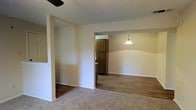 This single-story space is organized into a flowing open layout that connects the living area, dining space, and kitchen. The living area is finished with wall-to-wall carpet, providing a comfortable and quiet underfoot feel, while a ceiling fan overhead offers practical climate control. A partial wall divides the living area from the adjoining dining space, which transitions to hardwood-style flooring, creating a clear visual distinction between zones. A pendant light hangs above the dining area, providing focused overhead illumination and adding a subtle decorative touch. Beyond the dining area, the kitchen is visible and features dark wood cabinetry alongside a full-sized refrigerator, offering functional storage solutions. The walls throughout are finished in a neutral, light tone that helps reflect light and creates a clean, open feel. A textured popcorn ceiling runs throughout the space, consistent with a classic residential style. Overall, the layout is practical and straightforward, with clearly defined areas for living, dining, and cooking, making effective use of the available space on a single level.