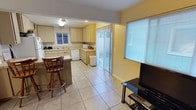 This single-floor space offers a functional and open layout that seamlessly connects the kitchen and living areas. The kitchen is outfitted with cream-toned cabinetry extending along two walls, providing ample storage and counter space. A tiled backsplash adds a clean, finished look, while white appliances, including a dishwasher and microwave, contribute to the bright, cohesive aesthetic. A peninsula with bar-height seating serves as both a casual dining spot and a natural divider between the kitchen and the adjacent living area. The flooring throughout is light-toned tile, lending a clean and easy-to-maintain surface across the entire space. Natural light enters generously through a window above the kitchen sink, a large slatted window in the living area, and a set of sliding glass doors fitted with vertical blinds, which provide direct access to an outdoor area. The living space accommodates a television setup, making it well-suited for everyday use. The overall ambiance is bright, practical, and straightforward, with a neutral color palette that keeps the space feeling open and welcoming.