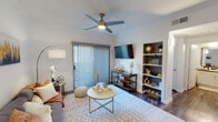 This single-story residence offers a thoughtfully designed layout that balances comfort and functionality across several distinct living areas. The living room serves as the heart of the home, featuring wood-style flooring, crown molding, and a contemporary ceiling fan with a built-in light fixture. A large sliding glass door fitted with vertical blinds floods the space with natural light and provides access to an outdoor area. Built-in shelving flanks the wall-mounted television, offering both display and storage space, while a patterned area rug anchors the seating arrangement.

Adjacent to the living room, a compact dining nook features two-tone gray walls with a chair rail detail and a large framed mirror that amplifies the sense of space. The galley-style kitchen is lined with white cabinetry, dark granite-style countertops, and wood-look flooring, providing ample storage and prep space alongside full-size appliances.

The primary bedroom features carpeted floors, a ceiling fan, and large windows dressed with sheer curtains that invite natural light. A second bedroom offers similar finishes with a bright window view. The bathroom includes a single vanity with dark countertop, a framed mirror, overhead cabinetry, and direct access to a spacious walk-in closet with built-in shelving and hanging rods.