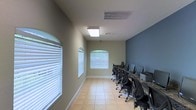 This single-floor computer lab is thoughtfully designed to support focused work and shared computing. The room features a long row of workstations lined against one wall, each equipped with desktop computers and ergonomic task chairs, providing ample space for multiple users to work simultaneously. The layout is clean and efficient, maximizing the available floor space for functional use.

The interior is finished with light ceramic tile flooring, which lends a bright and easy-to-maintain surface underfoot. Walls are painted in a neutral sage green on three sides, with a contrasting slate blue accent wall behind the workstations, adding a modern, professional touch to the environment. Arched windows fitted with white horizontal blinds allow natural light to filter into the space, contributing to a well-lit and welcoming atmosphere without glare on the screens. Overhead, a flush ceiling light fixture supplements the natural light, ensuring consistent illumination throughout the room. HVAC vents are integrated into the ceiling for year-round climate comfort. The combination of functional furnishings, natural light, and a clean color palette makes this computer lab a practical and pleasant environment for productivity.