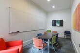 This single-floor professional space is thoughtfully designed to accommodate a variety of work styles, blending focused individual workstations with collaborative meeting areas. The workstation areas feature dark wood desks arranged with privacy dividers, providing each user with a dedicated, distraction-free environment. Colorful accent chairs in orange, yellow, and blue add a vibrant touch against the neutral carpeted flooring, which carries throughout the space. One workstation room is distinguished by a bold navy blue accent wall, creating a dynamic contrast to the soft, neutral tones found elsewhere. A framed abstract landscape painting with cool green and teal tones serves as a recurring decorative element, lending warmth and artistic character to the work areas. The collaborative meeting room is equipped with a large wall-mounted whiteboard, a conference-style table, and a mix of seating including a striking red upholstered sofa, making it well-suited for group discussions, presentations, or study sessions. Recessed lighting throughout ensures a well-lit, professional atmosphere. The overall layout is compact yet efficient, offering a complete suite of amenities for a productive and comfortable work environment.