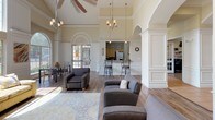Located at 125 S Murdeaux Ln in Dallas, Texas, this single-story community clubhouse offers a refined and welcoming environment designed for both relaxation and productivity. The main lounge area is an architectural highlight, boasting soaring vaulted ceilings, elegant crown molding, and dramatic arched windows that flood the space with natural light and frame views of the surrounding greenery. A classic tile-surround fireplace anchors one wall, complemented by built-in shelving and a wall-mounted TV, creating a comfortable gathering space. Warm hardwood-style flooring runs throughout, tying together the open layout. Seating areas are thoughtfully arranged with plush sofas, accent chairs, and decorative area rugs, while chandelier and ceiling fan fixtures add to the upscale ambiance. Graceful arched doorways and white wainscoting panels contribute to the space's traditional yet inviting character. A dedicated business center, enclosed by classic French doors, provides multiple computer workstations along built-in cabinetry and shelving, as well as a central table for collaborative use. Natural light enters through slatted window blinds, keeping the workspace bright. A kitchen bar area with counter seating and appliances supports community gatherings, while a separate dining area with table seating rounds out the functional amenities of this well-appointed clubhouse.