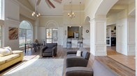 Located at 125 S Murdeaux Ln in Dallas, Texas, this single-story community clubhouse offers a refined and welcoming environment designed for both relaxation and productivity. The main lounge area is an architectural highlight, boasting soaring vaulted ceilings, elegant crown molding, and dramatic arched windows that flood the space with natural light and frame views of the surrounding greenery. A classic tile-surround fireplace anchors one wall, complemented by built-in shelving and a wall-mounted TV, creating a comfortable gathering space. Warm hardwood-style flooring runs throughout, tying together the open layout. Seating areas are thoughtfully arranged with plush sofas, accent chairs, and decorative area rugs, while chandelier and ceiling fan fixtures add to the upscale ambiance. Graceful arched doorways and white wainscoting panels contribute to the space's traditional yet inviting character. A dedicated business center, enclosed by classic French doors, provides multiple computer workstations along built-in cabinetry and shelving, as well as a central table for collaborative use. Natural light enters through slatted window blinds, keeping the workspace bright. A kitchen bar area with counter seating and appliances supports community gatherings, while a separate dining area with table seating rounds out the functional amenities of this well-appointed clubhouse.