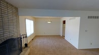 This two-story home offers a clean, functional layout with a welcoming main living area that sets the tone for the rest of the space. The living room is generously sized, featuring neutral-toned carpet that extends throughout, and is anchored by a brick fireplace with classic iron accessories — a standout architectural detail that adds warmth and character to the room. A large window with vertical blinds allows natural light to filter in, brightening the open space. A pendant light fixture hangs centrally from the ceiling, providing both ambient lighting and a subtle decorative touch. The walls are finished in crisp white, contributing to a bright and airy atmosphere throughout. A doorway visible from the main living area provides a seamless transition to adjacent spaces, including what appears to be a kitchen with appliances beyond. Vents integrated into the walls indicate a central HVAC system for year-round climate comfort. The overall design is straightforward and practical, with a neutral palette that lends itself well to a variety of furnishing styles. Across both floors, the home presents a solid foundation of classic residential architecture with key functional features in place.