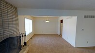 This two-story home offers a clean, functional layout with a welcoming main living area that sets the tone for the rest of the space. The living room is generously sized, featuring neutral-toned carpet that extends throughout, and is anchored by a brick fireplace with classic iron accessories — a standout architectural detail that adds warmth and character to the room. A large window with vertical blinds allows natural light to filter in, brightening the open space. A pendant light fixture hangs centrally from the ceiling, providing both ambient lighting and a subtle decorative touch. The walls are finished in crisp white, contributing to a bright and airy atmosphere throughout. A doorway visible from the main living area provides a seamless transition to adjacent spaces, including what appears to be a kitchen with appliances beyond. Vents integrated into the walls indicate a central HVAC system for year-round climate comfort. The overall design is straightforward and practical, with a neutral palette that lends itself well to a variety of furnishing styles. Across both floors, the home presents a solid foundation of classic residential architecture with key functional features in place.