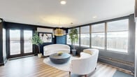 This sophisticated single-floor space makes a striking first impression with its deep navy-painted walls contrasted against light hardwood flooring, creating a bold yet inviting atmosphere. The main lounge area is a standout, anchored by curved white sofas and a sculptural round black coffee table arranged atop a woven area rug. A large gold drum pendant light hangs overhead, adding a warm, upscale focal point to the room. Lush indoor trees are positioned throughout, bringing a refreshing natural element to the modern interior. Expansive floor-to-ceiling windows wrap around the lounge, flooding the space with natural light and offering sweeping panoramic views of the surrounding cityscape. A secondary seating area features channel-tufted bench seating paired with a dark wood table, illuminated by globe pendant fixtures — ideal for meetings or collaborative gatherings. The corridor connecting the areas is lined with artwork and directional track lighting, maintaining the space's curated aesthetic. A wall-mounted split AC unit and baseboard heating ensure year-round comfort. An exterior door provides direct outdoor access, adding flexibility to the space's functionality. Overall, this space exudes a contemporary, design-forward ambiance suited for professional or social use.