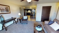This single-floor residential unit offers a thoughtfully designed open-concept layout that connects the living, dining, and kitchen areas in one cohesive space. The living area features plush carpeted flooring and is furnished with comfortable seating, including a patterned accent chair and a sofa, complemented by side tables and decorative accents that add a warm, inviting ambiance. A ceiling fan overhead provides both style and function.

The space transitions naturally into the dining area, where a bistro-style table and chairs create an intimate setting for meals. Dark wood tones in the furniture contrast tastefully with the lighter walls and flooring throughout.

The kitchen is well-equipped with dark cabinetry, modern stainless steel appliances including a refrigerator and microwave, and a classic tile backsplash. Pendant lighting above adds a refined touch to the cooking and preparation area.

The unit also features multiple interior doors leading to additional private spaces, finished with rich dark wood tones that carry consistently throughout the unit's design. The overall aesthetic is clean, modern, and functional, with a neutral color palette that creates a bright and welcoming atmosphere ideal for everyday living.