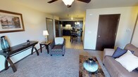 This single-floor residential unit offers a thoughtfully designed open-concept layout that connects the living, dining, and kitchen areas in one cohesive space. The living area features plush carpeted flooring and is furnished with comfortable seating, including a patterned accent chair and a sofa, complemented by side tables and decorative accents that add a warm, inviting ambiance. A ceiling fan overhead provides both style and function.

The space transitions naturally into the dining area, where a bistro-style table and chairs create an intimate setting for meals. Dark wood tones in the furniture contrast tastefully with the lighter walls and flooring throughout.

The kitchen is well-equipped with dark cabinetry, modern stainless steel appliances including a refrigerator and microwave, and a classic tile backsplash. Pendant lighting above adds a refined touch to the cooking and preparation area.

The unit also features multiple interior doors leading to additional private spaces, finished with rich dark wood tones that carry consistently throughout the unit's design. The overall aesthetic is clean, modern, and functional, with a neutral color palette that creates a bright and welcoming atmosphere ideal for everyday living.