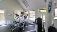 This single-floor fitness center is thoughtfully designed to support a well-rounded workout routine. The room features a vaulted ceiling that lends an open, airy feel to the space, enhancing the sense of roominess despite its intimate footprint. Overhead, recessed lighting fixtures complement the abundant natural light that pours in through multiple wood-framed windows lining the walls, offering pleasant views of the surrounding outdoor greenery.

The fitness center is equipped with a solid selection of cardio and strength training machines. Cardio equipment includes treadmills, an elliptical trainer, and a stationary bike — with recognizable brands such as Precor and Vectra present. A multi-station strength training machine rounds out the offerings, providing options for resistance-based workouts. A wall-mounted mirror on one side of the room enhances the sense of space while allowing users to monitor their form during exercise.

The flooring is a durable, dark rubberized surface well-suited for a fitness environment. The neutral, light-gray wall tones keep the atmosphere clean and focused. A wall-mounted television is also present, offering entertainment during workouts. Overall, the space delivers a practical and comfortable environment for fitness enthusiasts.