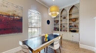 Located at 13100 W Bellfort Ave in Houston, TX, this thoughtfully designed single-floor space combines a dining room and a dedicated office area into a cohesive, functional layout. Light wood-tone flooring runs throughout, lending warmth and continuity between the two areas.

The dining room is an inviting space anchored by a substantial dark-topped dining table with mid-century modern chairs upholstered in white. A tiered gold pendant light hangs overhead, casting a warm glow over the room. An arched window with slatted blinds allows natural light to filter in, while crown molding and built-in shelving with elegant arched detailing add refined architectural character. The shelves display books, decorative objects, and cabinetry below for ample storage.

The adjacent office area is practical and well-equipped, featuring an L-shaped wraparound desk with multiple workstations and monitors, accommodating collaborative or multi-user work. White upholstered chairs provide comfortable seating, and decorative wall art contributes a creative touch to the professional setting. The arched architectural motif from the dining area carries through, reinforcing a consistent design language across the space. Together, both areas create an elegant, functional environment well-suited for meetings, work, and refined gatherings.