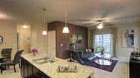 This single-floor residence showcases a thoughtfully designed open-concept layout that seamlessly connects the kitchen and living areas. The kitchen serves as a central hub, featuring granite countertops, a double stainless steel sink, and dark cabinetry. A breakfast bar peninsula with elegant pendant lighting creates a natural transition into the living space, making it ideal for entertaining.

The living area is anchored by a bold, deep plum accent wall that adds warmth and visual depth to the space. A ceiling fan with integrated lighting provides both comfort and ambiance. The room is furnished with a contemporary sofa adorned with decorative pillows, complemented by a sleek glass coffee table and a stylish accent chair.

Large sliding glass doors at the far end of the living area flood the space with natural light and offer views of the outdoors, creating an airy and open atmosphere. Neutral wall tones throughout the main areas contrast beautifully with the dramatic accent wall, lending the space a balanced and inviting character. Decorative touches such as framed artwork, fresh floral arrangements, and curated accessories complete the polished, move-in-ready aesthetic of this well-appointed home.