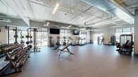 Located at 360 S Rio Grande Street in Salt Lake City, Utah, this well-appointed fitness center offers a generously sized, open-plan layout across a single floor. The space is defined by its industrial-modern aesthetic, featuring exposed ductwork and piping along the ceiling, sleek linear LED lighting, and polished concrete-style ceilings that lend the room a contemporary, loft-like character. Large floor-to-ceiling windows line the perimeter walls, flooding the interior with abundant natural light and providing open views of the surrounding neighborhood.

The durable rubber flooring throughout is well-suited for high-traffic exercise use and provides a safe, cushioned surface across the entire workout area. The cardio zone is equipped with multiple treadmills, elliptical trainers, and stationary bikes, each featuring individual display screens, with wall-mounted flat-screen TVs positioned for visibility from the equipment. The strength training area includes a cable cross machine, a leg press machine, adjustable benches, a dumbbell rack, and additional resistance machines, offering a comprehensive range of workout options.

A large ceiling fan provides additional air circulation, while full-length mirrors along one wall enhance the sense of space. The fitness center is accessible via keypad-secured entry doors, ensuring a controlled and secure environment for users.