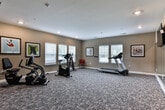 Located at 170 SR-113 in Carrollton, Georgia, this single-floor fitness center offers a well-appointed space designed to support a range of workout routines. The room features a neutral warm beige color palette on the walls, complemented by white trim and a textured, patterned carpet that runs throughout the entire floor area.

The main exercise zone is equipped with cardio machines, including a treadmill, upright stationary bike, recumbent bike, and an elliptical trainer, providing versatile options for cardiovascular workouts. A large wall-mounted mirror spans one side of the room, enhancing the sense of space and allowing users to monitor their form during exercise. A flat-screen television is mounted adjacent to the mirror for entertainment during workouts.

Natural light filters through multiple white-slatted blinds covering several windows, creating a bright and inviting atmosphere. Recessed ceiling lighting provides consistent illumination throughout. Framed decorative artwork, including whimsical dog portraits and bicycle-themed prints, adds a welcoming, lighthearted character to the space. Additional fitness accessories, such as a stability ball, are also available. A dedicated entry door and posted fitness center guidelines round out the functional, community-oriented design of the facility.