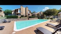 This single-story community showcases a well-appointed outdoor pool area as its central amenity. The generously sized rectangular swimming pool features clear, turquoise water and is framed by a broad concrete deck furnished with multiple reclining lounge chairs, ideal for sunbathing. Terraced stone retaining walls border one side of the pool, adding a structured, natural aesthetic to the landscaping while separating the pool level from the surrounding grounds.

The adjacent residential buildings display a warm stucco exterior in earthy tan tones, accented by prominent exterior chimneys and private balconies that overlook the pool area. Mature trees and well-maintained shrubs surround the perimeter, providing shade and a sense of privacy. A chain-link fence encloses the pool area, densely lined with climbing vines and leafy hedges that create a natural green screen.

A secluded patio space is also present, featuring a round table and chairs nestled among lush, overgrown greenery, offering a quiet retreat. The overall ambiance is relaxed and residential, combining functional outdoor amenities with established landscaping that contributes to a comfortable, community-oriented environment.