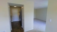 This single-floor space is organized with distinct functional areas separated by a defining wall. The living area features neutral-toned carpet flooring, contributing to a clean, understated aesthetic, while the adjoining kitchen and entryway transition to dark hardwood-style flooring, creating a clear visual boundary between spaces. The kitchen is equipped with a stainless steel refrigerator, and a doorway visible from the living area suggests an additional room or closet beyond. Walls throughout are finished in a soft, neutral tone, complementing the light-colored trim and baseboards. The layout incorporates a flush ceiling light in the kitchen/hallway area, providing functional illumination. The overall design is straightforward and practical, with an open sightline between the living space and kitchen corridor that enhances the sense of flow within the unit. The clean, move-in-ready condition of the space is evident, with minimal ornamentation allowing the structural layout and flooring details to stand out as the primary defining characteristics.