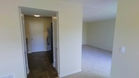 This single-floor space is organized with distinct functional areas separated by a defining wall. The living area features neutral-toned carpet flooring, contributing to a clean, understated aesthetic, while the adjoining kitchen and entryway transition to dark hardwood-style flooring, creating a clear visual boundary between spaces. The kitchen is equipped with a stainless steel refrigerator, and a doorway visible from the living area suggests an additional room or closet beyond. Walls throughout are finished in a soft, neutral tone, complementing the light-colored trim and baseboards. The layout incorporates a flush ceiling light in the kitchen/hallway area, providing functional illumination. The overall design is straightforward and practical, with an open sightline between the living space and kitchen corridor that enhances the sense of flow within the unit. The clean, move-in-ready condition of the space is evident, with minimal ornamentation allowing the structural layout and flooring details to stand out as the primary defining characteristics.