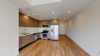 This single-floor unit showcases a thoughtfully designed, modern interior with a cohesive aesthetic carried throughout every room. The open-concept kitchen is a standout feature, outfitted with flat-front wood-grain cabinetry extending from floor to ceiling, sleek white countertops, and a full suite of stainless steel appliances including a gas range, built-in microwave, dishwasher, and a large side-by-side refrigerator with an in-door water and ice dispenser. Recessed lighting and a skylight element above the kitchen provide excellent illumination. Light-toned hardwood-style flooring flows seamlessly across the entire unit, creating a sense of visual continuity and warmth. The bedrooms are bright and generously proportioned, with crisp white walls, and at least one room features a wide window with outdoor views and another includes a sliding-door closet. Two bathrooms serve the unit — one with a glass-enclosed walk-in shower and wood-tone vanity cabinetry, and another with a full bathtub and glass sliding doors, each finished with white countertops and large mirrors. The overall ambiance is clean, contemporary, and move-in ready, with a neutral palette that allows for versatile interior styling.