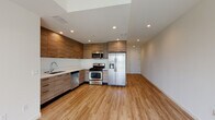 This single-floor unit showcases a thoughtfully designed, modern interior with a cohesive aesthetic carried throughout every room. The open-concept kitchen is a standout feature, outfitted with flat-front wood-grain cabinetry extending from floor to ceiling, sleek white countertops, and a full suite of stainless steel appliances including a gas range, built-in microwave, dishwasher, and a large side-by-side refrigerator with an in-door water and ice dispenser. Recessed lighting and a skylight element above the kitchen provide excellent illumination. Light-toned hardwood-style flooring flows seamlessly across the entire unit, creating a sense of visual continuity and warmth. The bedrooms are bright and generously proportioned, with crisp white walls, and at least one room features a wide window with outdoor views and another includes a sliding-door closet. Two bathrooms serve the unit — one with a glass-enclosed walk-in shower and wood-tone vanity cabinetry, and another with a full bathtub and glass sliding doors, each finished with white countertops and large mirrors. The overall ambiance is clean, contemporary, and move-in ready, with a neutral palette that allows for versatile interior styling.