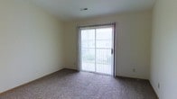 Located at 150 Reed Way in Sunburn, Ohio, this single-floor space offers a straightforward and functional layout. The interior features wall-to-wall carpeting that adds warmth and comfort underfoot, complemented by neutral-toned walls that create a clean, versatile backdrop suitable for a variety of uses. A standout feature of the main living area is the full-width sliding glass door, dressed with vertical blinds, which provides direct access to the outdoors and invites an abundance of natural light into the space. The view through the glass door reveals a green outdoor setting, enhancing the sense of openness and connection to the surrounding environment. A ceiling-mounted HVAC vent ensures year-round climate control throughout the interior. The overall ambiance is bright, understated, and move-in ready, offering a comfortable and practical environment within a single, open-concept level.