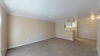 This two-story residence offers a practical and comfortable layout across two well-defined levels, connected by a carpeted staircase. The main level features a generously sized living area finished with warm beige walls, plush carpet flooring, and elegant crown molding that frames the space. A pass-through kitchen counter opens directly to the living area, providing a functional connection between the two spaces. White cabinetry and overhead ventilation are visible in the kitchen, lending a clean, organized aesthetic.

The upper level hosts a spacious bedroom retreat complete with a ceiling fan and light fixture, a closet, and direct access to a full en-suite bathroom. The bathroom features a single-sink vanity with a light countertop, white cabinetry with storage drawers, a large mirror, and a combined shower and bathtub with hardwood-style flooring.

An additional carpeted bedroom on the lower level offers crown molding, a chair rail detail along the walls, and a ceiling-mounted light fixture, providing a well-finished and cozy feel. Throughout the residence, consistent warm neutral tones, white trim, and carpeted floors create a cohesive and inviting atmosphere. The upper bedroom benefits from natural light through a window with blinds, adding brightness to the space.