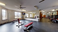 Located at 9815 Seasons West Dr. in Indianapolis, Indiana, this two-story fitness center is a comprehensively appointed facility designed to support a full range of workout routines. The main level features generously sized exercise areas finished with durable rubber flooring throughout. Large wall-mounted mirrors and multiple windows allow natural light to fill the space, creating an open and motivating environment. Ceiling fans provide added comfort across the workout zones.

Strength training areas are equipped with weight benches, barbells, and multiple dumbbell racks offering a wide range of weight options, complemented by storage cubbies for personal belongings. A dedicated cardio section houses rowing machines, recumbent bikes, and additional cardio equipment positioned alongside floor-to-ceiling windows that offer outdoor views of the surrounding community.

A striking spiral staircase connects the two levels, adding an architectural focal point to the space. The upper floor features elliptical trainers, treadmills, and additional strength machines, along with a windowed interior overlook into adjacent common areas framed by rich wood trim and stone accent walls. The upper level also benefits from soaring windows that flood the space with natural light. Throughout, the facility maintains a clean, functional, and well-organized aesthetic suited for everyday fitness use.