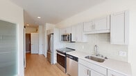This single-floor unit presents a clean, modern aesthetic throughout its well-designed layout. The kitchen serves as a functional centerpiece, outfitted with a full suite of stainless steel appliances, including a refrigerator, electric range, built-in microwave, and dishwasher. Crisp white shaker-style cabinetry extends from floor to ceiling, offering generous storage, while light quartz countertops and a classic white subway tile backsplash contribute to the kitchen's bright, contemporary character. Light-toned hardwood-style flooring runs throughout the kitchen and main living areas, enhancing the sense of openness. A window in the kitchen area admits natural light and provides views of surrounding greenery.

The bedrooms feature neutral carpet flooring and are finished in soft, warm tones. Each room includes closet space equipped with wire shelving systems, offering practical storage solutions. One bedroom features a walk-in style closet configuration, while another includes a sliding-door closet.

The bathroom is finished with a single-sink vanity topped with a gray countertop, a large mirror with modern bar lighting above, and a glass-door shower-tub combination. Light hardwood-style flooring carries through the bathroom, tying the space together with a cohesive, polished feel throughout.