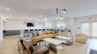 This single-floor communal space offers a generously sized, open-plan layout finished with light wood-look flooring and crisp white walls that create a bright, welcoming atmosphere throughout. The interior is thoughtfully divided into distinct functional zones that flow seamlessly into one another.

The main lounge area serves as the focal point, furnished with a large tan sectional sofa, gold-framed leather accent chairs, and paired white coffee tables arranged atop a neutral area rug. A sculptural multi-arm pendant light anchors the seating arrangement from above, complemented by recessed ceiling lighting throughout.

A standout feature wall adorned with an abstract mural in soft blush and cream tones provides a striking backdrop for a wall-mounted television and media console, adding an artistic element to the space.

Toward the rear, a wet bar with dark cabinetry and a white countertop is accompanied by bar-height seating, while a separate dining zone features tall bistro tables with black chairs positioned beneath large windows that allow ample natural light to filter in. A geometric cage-style pendant light fixture illuminates this area.

The space also includes a long communal bar counter with additional bar stools, ideal for collaborative or casual use, reinforcing the space's versatile, social character.