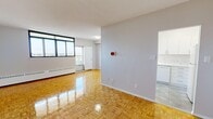 This 2-bedroom, 1-bathroom unit is laid out efficiently across a single floor, offering a practical and well-organized living space. Polished parquet flooring flows consistently throughout the living areas and bedrooms, lending a warm, classic character to the interior. The living room is a generous, open space anchored by wide, horizontal windows that invite abundant natural light and offer open views of the surrounding landscape, creating a bright and airy atmosphere. Baseboard heaters line the perimeter, providing consistent warmth throughout the unit. The kitchen is cleanly finished with white cabinetry, white appliances including an electric range, and a marble-style tile backsplash that adds a refined touch. Light grey tile flooring distinguishes the kitchen from the rest of the unit. Both bedrooms continue the parquet flooring theme; the primary bedroom features a large sliding-door closet offering ample built-in storage, while the second bedroom includes a window with open outdoor views. The bathroom is finished with floor-to-ceiling marble-style tile surrounding a full soaking tub with overhead shower. A hallway connects the rooms cohesively, completing this functional and well-appointed residential unit.