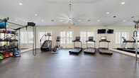 This fitness center is a comprehensively equipped facility designed to support a full range of workout routines. The space features a clean, modern aesthetic with white walls, recessed lighting, and a large ceiling fan that contribute to a bright and airy atmosphere. Dark rubberized flooring spans the entire area, providing durability and cushioning underfoot.

The cardio zone is prominently positioned near a bank of French doors and large windows fitted with white plantation shutters, allowing ample natural light to fill the room. Equipment in this area includes multiple treadmills with individual screens, a stair climber, and an elliptical trainer. A wall-mounted flat-screen television is positioned for easy viewing during cardio sessions.

An adjacent area houses a dedicated strength and free-weight zone, featuring a multi-tiered dumbbell rack stocked with a wide range of weights, medicine balls, stability balls, and a multi-station cable and rack system. Weight benches are available for a variety of exercises.

On the opposite side of the space, a cycling area offers spin bikes alongside low-profile rowing or sliding machines, providing additional low-impact cardio options. A mirrored wall in the weight area enhances the sense of space and supports proper form during training. The overall environment is clean, organized, and well-maintained.
