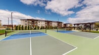 Located at 780 Doss Dr in Marysville, Ohio, this outdoor recreational court complex offers a well-appointed multi-court facility designed for both pickleball and tennis play. The courts feature a distinctive two-tone surface with green outer zones and bright blue infield sections, clearly marked with crisp white lines and equipped with regulation nets. Tall light poles are strategically positioned throughout the facility, enabling play during evening hours. The complex is fully enclosed by a black chain-link fence, providing both safety and boundary definition. A neatly maintained row of columnar evergreen trees lines the perimeter, adding a touch of greenery and serving as a natural windbreak and visual screen. A pergola structure is visible adjacent to the courts, offering a shaded gathering area for players and spectators. The surrounding landscape features manicured lawns and open skies that create an inviting, open-air atmosphere. Multi-story residential buildings are visible in the background, indicating that this facility serves as an amenity within a broader residential community. The courts are well-maintained and thoughtfully designed to accommodate multiple players simultaneously, making it an ideal space for both casual recreation and organized play.